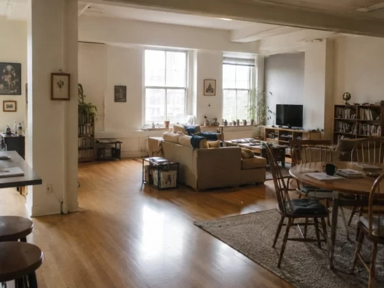A spacious 1990s-style loft apartment with warm wood floors, large windows, subtle industrial features, and naturally divided living and dining areas with lived-in furniture