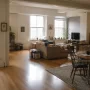 A spacious 1990s-style loft apartment with warm wood floors, large windows, subtle industrial features, and naturally divided living and dining areas with lived-in furniture